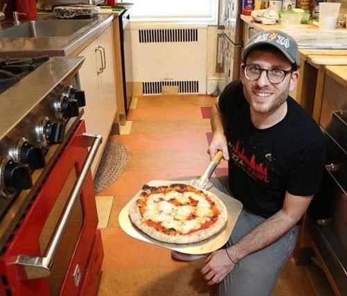 A photo of a smiling man kneeling next to an oven with a pizza